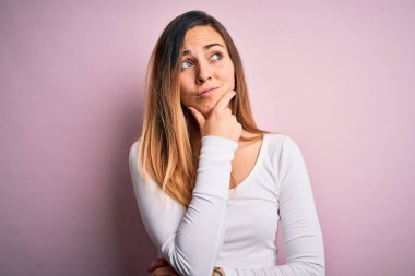 Pembe arka planda beyaz tişört giyen, mavi gözlü genç, güzel sarışın kadın bir soru hakkında endişeli ve gergin bir şekilde düşünüyor.