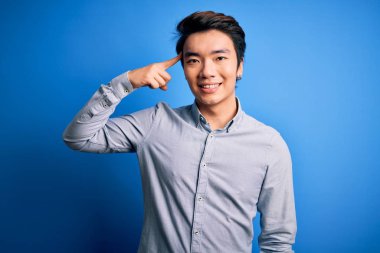 Genç, yakışıklı, sıradan bir gömlek giyen, mavi arka planda duran, tek parmağıyla gülümseyen, harika bir fikir ya da düşünce, iyi bir hafıza.