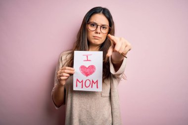 Elinde sevgi dolu bir kağıt tutan genç bir kadın anneler gününü kamerayı işaret ederek kutluyor ve sen, el işareti, ön taraftan pozitif ve kendinden emin bir jest.