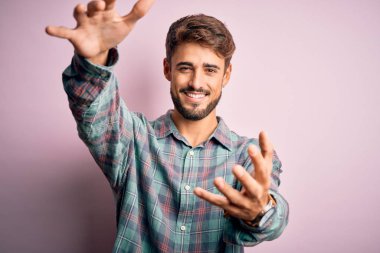 Sakallı, sıradan gömlekli, pembe arka planda dikilmiş kameraya sarılmak için gülümseyen genç yakışıklı bir adam. Mutluluğu kucaklayan neşeli bir ifade..