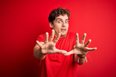 Kıvırcık saçlı, kırmızı arka planda sıradan tişörtler giyen, korku ifadesinden korkan ve dehşete düşmüş genç sarışın adam. El hareketi yapmayı kes, şok içinde bağır. Panik konsepti.