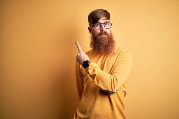 Handsome Irish redhead man with beard wearing glasses over yellow isolated background Pointing with hand finger to the side showing advertisement, serious and calm face