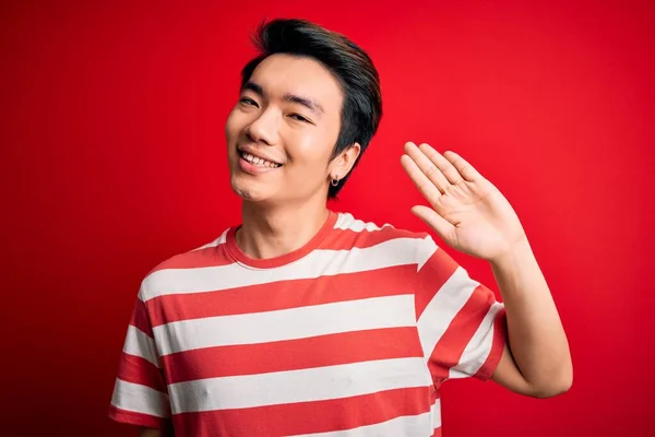 若いハンサムな中国人男性を着てカジュアルな縞模様のTシャツ赤い背景の上に立って手を振っこんにちは幸せと笑顔 優しい歓迎ジェスチャー — ストック写真