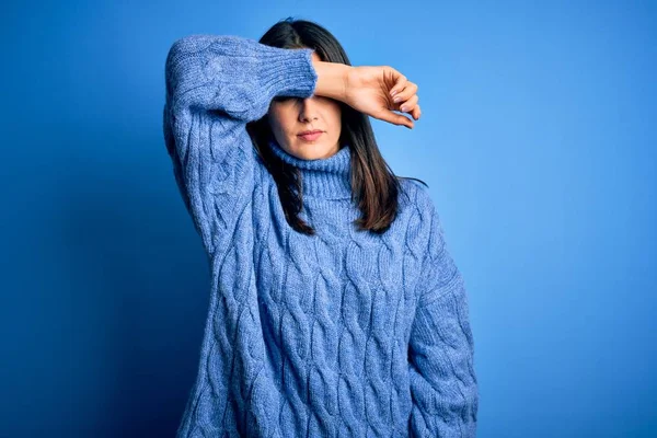 Young brunette woman with blue eyes wearing casual turtleneck sweater covering eyes with arm, looking serious and sad. Sightless, hiding and rejection concept