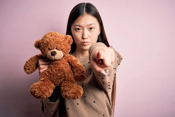 Young asian woman hugging teddy bear stuffed animal over pink isolated background pointing with finger to the camera and to you, hand sign, positive and confident gesture from the front