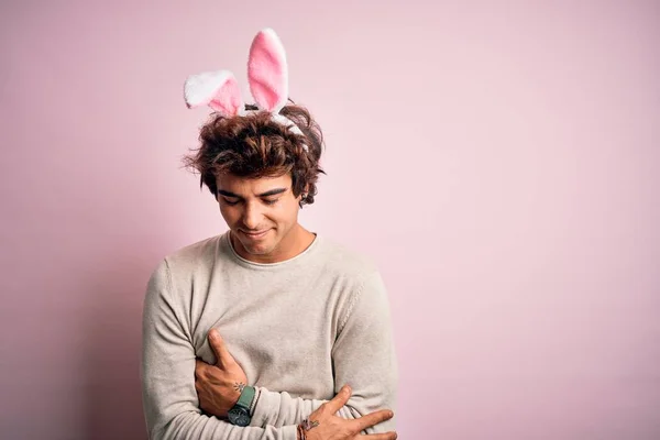 年轻英俊的男子抱着东方兔子的耳朵 手托着肚子站在孤立的粉色背景上 因为消化不良 痛苦不堪 阿切概念 — 图库照片