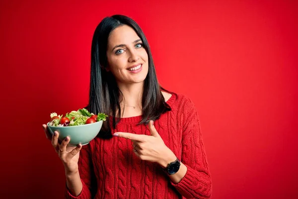 若いですブルネット女性とともに青い目食べる健康的な緑のサラダ以上隔離された赤背景非常に幸せなポインティングとともに手と指 — ストック写真