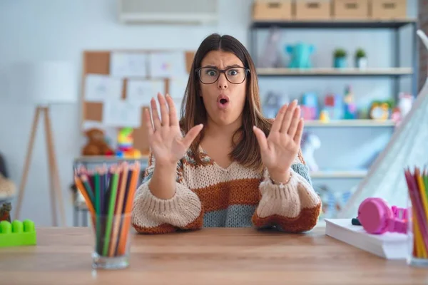 Young beautiful teacher woman wearing sweater and glasses sitting on desk at kindergarten Moving away hands palms showing refusal and denial with afraid and disgusting expression. Stop and forbidden.