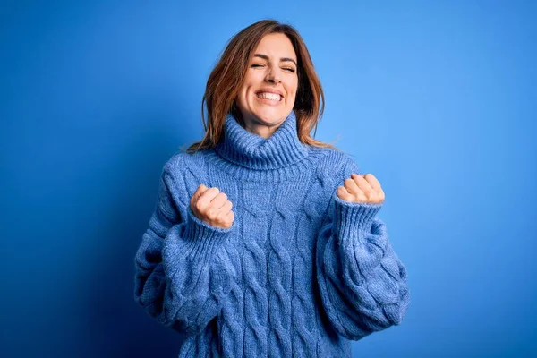 Mavi arka plan üzerine balıkçı yaka kazak giyen genç ve güzel bir kadın. Kolları havada ve gözleri kapalı zaferi kutlarken gülümsüyor. Kazanan konsept.