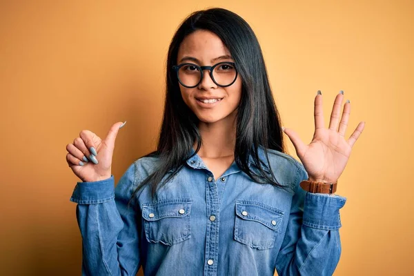 Young beautiful chinese woman wearing casual denim shirt over isolated yellow background showing and pointing up with fingers number six while smiling confident and happy.