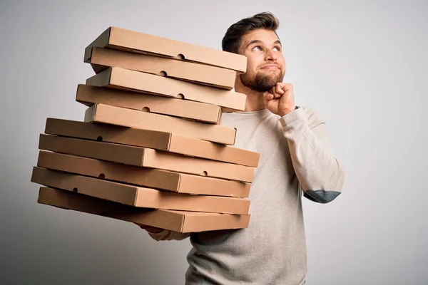 Young blond man with beard and blue eyes holding cardboards of pizza over white background serious face thinking about question, very confused idea