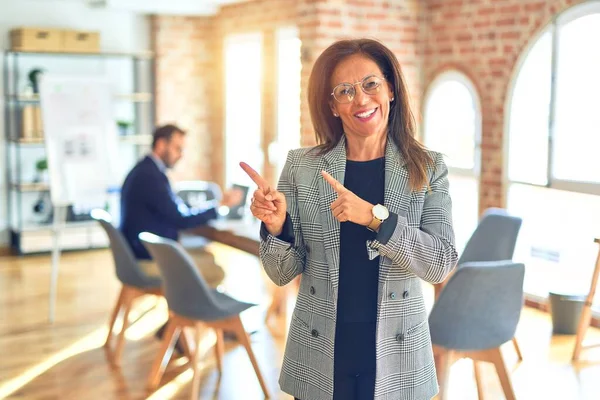 Middle age beautiful businesswoman wearing jacket and glasses standing at the office smiling and looking at the camera pointing with two hands and fingers to the side.