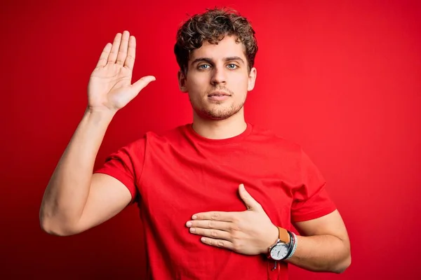 Young blond handsome man with curly hair wearing casual t-shirt over red background Swearing with hand on chest and open palm, making a loyalty promise oath
