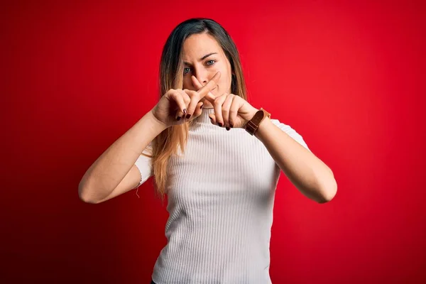 Beautiful blonde woman with blue eyes wearing casual white t-shirt over red background Rejection expression crossing fingers doing negative sign