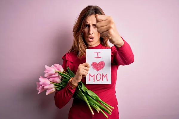 Beautiful brunette woman holding love mom message and tulips celebrating mothers day annoyed and frustrated shouting with anger, crazy and yelling with raised hand, anger concept