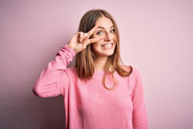 Genç, güzel, kızıl saçlı bir kadın pembe arka planda günlük süveter giyiyor. Barış sembolü yapıyor. Yüzünde parmaklar, yüzünde neşeyle zafer işareti yapıyor.