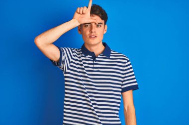 Genç bir çocuk mavi izole edilmiş arka planda dikilmiş tişört giyiyor. Alnında parmakları olan insanlarla alay ediyor. Ezik hareketler yapıyor, alay ediyor..