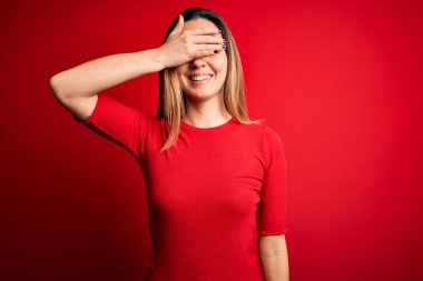Kırmızı arka planda sıradan tişörtler giyen mavi gözlü genç, güzel, sarışın bir kadın. Gülümsüyor ve yüz yüze gülüyor. Şaşırtmak için gözleri örtüyor. Kör kavram.