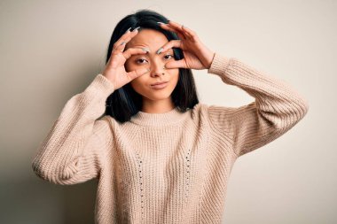 İzole edilmiş beyaz arka plan üzerine günlük süveter giyen genç, güzel Çinli kadın sabah yorgunluğu yüzünden uykulu ve yorgun bir şekilde gözlerini açmaya çalışıyor.