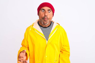 Yağmurluk giyen ve yünlü şapka takan orta yaşlı bir adam izole edilmiş beyaz arka planda durup dudaklı balık suratı yapıyor, çılgınca ve komik bir jest. Komik ifade.