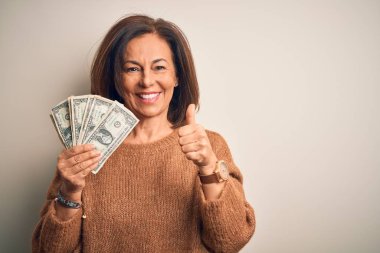 Orta yaşlı esmer kadın, elinde bir dolar banknotla izole edilmiş arka planda mutlu, gülümseyen, işaret veren, parmaklarıyla başparmağını kaldıran, mükemmel bir işaret.
