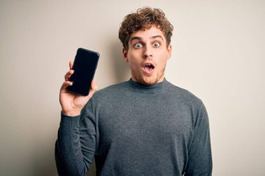 Sarışın, kıvırcık saçlı, akıllı telefon gösteren genç bir adam izole edilmiş beyaz arka planda duruyor. Şaşırmış bir yüzle korkmuş ve korku ifadesinden heyecanlanmış.