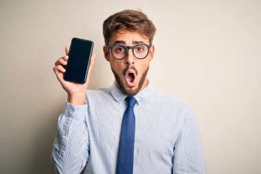 Akıllı telefonu elinde tutan genç bir işadamı, izole edilmiş beyaz arka planda duruyor. Şaşırmış bir yüzle korkmuş ve korku ifadesinden heyecanlanmış bir halde.