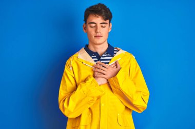 Sarı yağmurluk giyen genç balıkçı çocuk izole edilmiş arka planda gülümsüyor elleri göğsünde, gözleri kapalı ve yüzünde minnet dolu bir jest var. Sağlık kavramı.