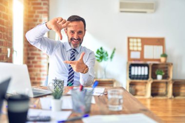 Middle age handsome businessman wearing tie sitting using laptop at the office smiling making frame with hands and fingers with happy face. Creativity and photography concept.