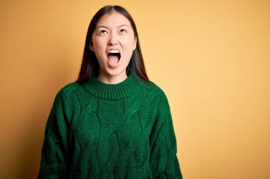 Sarı arka plan üzerine yeşil kazak giyen genç ve güzel Asyalı kadın öfkeli ve öfkeli çığlıklar atıyor, öfkeyle bağırıyor. Öfke ve agresif kavram.