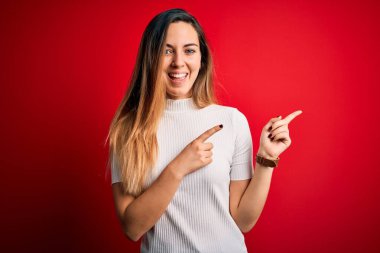 Mavi gözlü, güzel, beyaz tişörtlü, kırmızı arka planda gülümseyen ve kameraya bakan iki eliyle ve parmaklarıyla yüzünü gösteren bir kadın..