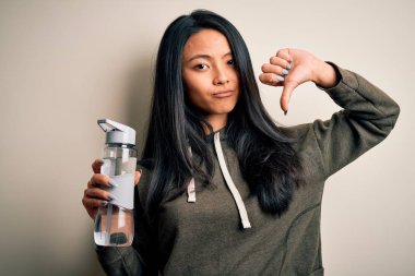 Genç, güzel Çinli sporcu kadın izole edilmiş beyaz arka planda su şişesi tutuyor. Kızgın bir yüzü var. Negatif işaretler baş parmakları aşağıda, reddedilme kavramından hoşlanmadığını gösteriyor.