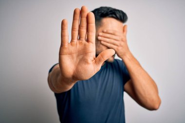 Genç ve yakışıklı bir adam, günlük tişört giyip izole edilmiş beyaz arka planda dikiliyor, gözleri ellerle örtüyor ve üzgün ve korku ifadeleriyle jest yapıyor. Utanç verici ve olumsuz bir kavram..