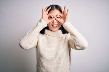 Young beautiful asian woman wearing casual sweater standing over isolated background doing ok gesture like binoculars sticking tongue out, eyes looking through fingers. Crazy expression.