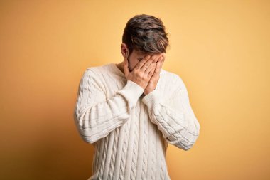 Sarışın, sakallı ve mavi gözlü genç adam sarı arka planda beyaz kazak giyiyor ve ağlarken suratını ellerle kapatıyor. Depresyon kavramı.