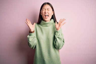 Young beautiful asian woman wearing green winter sweater over pink solated background celebrating mad and crazy for success with arms raised and closed eyes screaming excited. Winner concept