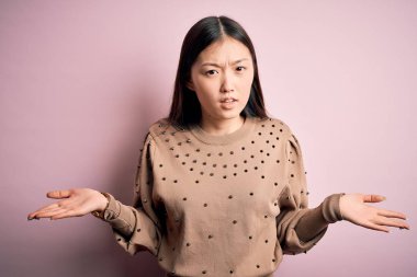 Young beautiful asian woman wearing fashion and elegant sweater over pink solated background clueless and confused with open arms, no idea concept.