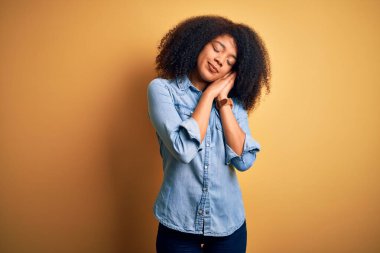 Sarıya boyanmış arka planda duran Afro saçlı, genç ve güzel Afro-Amerikalı kadın yorgun uyur ve elleri ve elleri kapalı gülümserken poz verir..