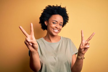 Kıvırcık saçlı, sıradan tişörtlü, kameraya bakıp zafer işareti yapan güzel Afro-Amerikan kadın. İki numara..