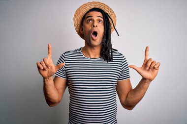 Afrika kökenli Amerikalı genç bir adam tatilde. Çizgili tişört ve şapka giyiyor. Şaşkın şaşkın yukarı bakıyor, parmaklarıyla işaret ediyor ve kollarını kaldırıyor..