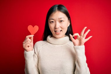 Young asian woman holding romantic red heart paper shape over red isolated background doing ok sign with fingers, excellent symbol