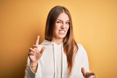 Young beautiful redhead sporty woman wearing sweatshirt over isolated yellow background disgusted expression, displeased and fearful doing disgust face because aversion reaction. With hands raised