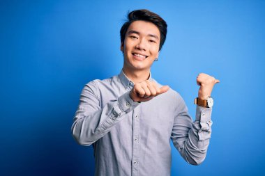 Genç, yakışıklı, sıradan bir gömlek giyen, mavi arka planda duran, elleri ve başparmakları havada, kendine güvenen, sırıtan Çinli bir adam.