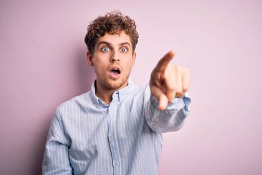 Sarışın, kıvırcık saçlı, beyaz arka planda çizgili tişörtlü, genç ve yakışıklı bir adam. Parmakları önde, ağzı açık, şaşkın, ön tarafta bir şey.