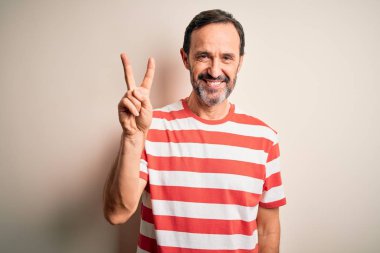 Orta yaşlı, beyaz çizgili tişört giyen, izole edilmiş beyaz arka plan üzerinde duran, iki numarayı işaret eden, kendinden emin ve mutlu gülümseyen, orta yaşlı adam..
