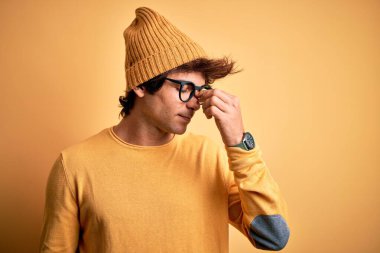 İzole edilmiş sarı arka plan üzerinde gündelik tişört ve gözlük takan genç yakışıklı adam yorgunluk ve baş ağrısı hissederek burnunu ve gözlerini ovuşturuyor. Stres ve hüsran kavramı.