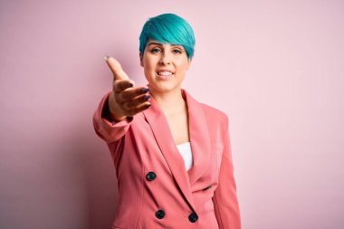 Young beautiful businesswoman with blue fashion hair wearing jacket over pink background smiling friendly offering handshake as greeting and welcoming. Successful business.