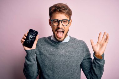 Sakallı, gözlüklü, kırık ve çatırdayan akıllı telefonlu genç adam çok mutlu ve heyecanlı, kazanan yüz ifadesi zaferi büyük bir gülümsemeyle kutluyor ve ellerini kaldırıyor.
