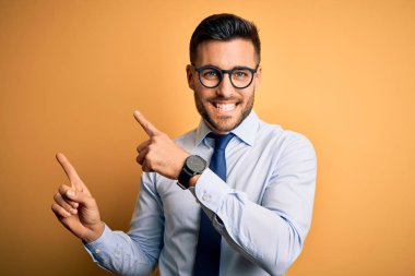 Young handsome businessman wearing tie and glasses standing over yellow background smiling and looking at the camera pointing with two hands and fingers to the side.