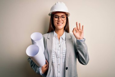 Young beautiful architect woman wearing safety helmet and glasses holding blueprints doing ok sign with fingers, excellent symbol